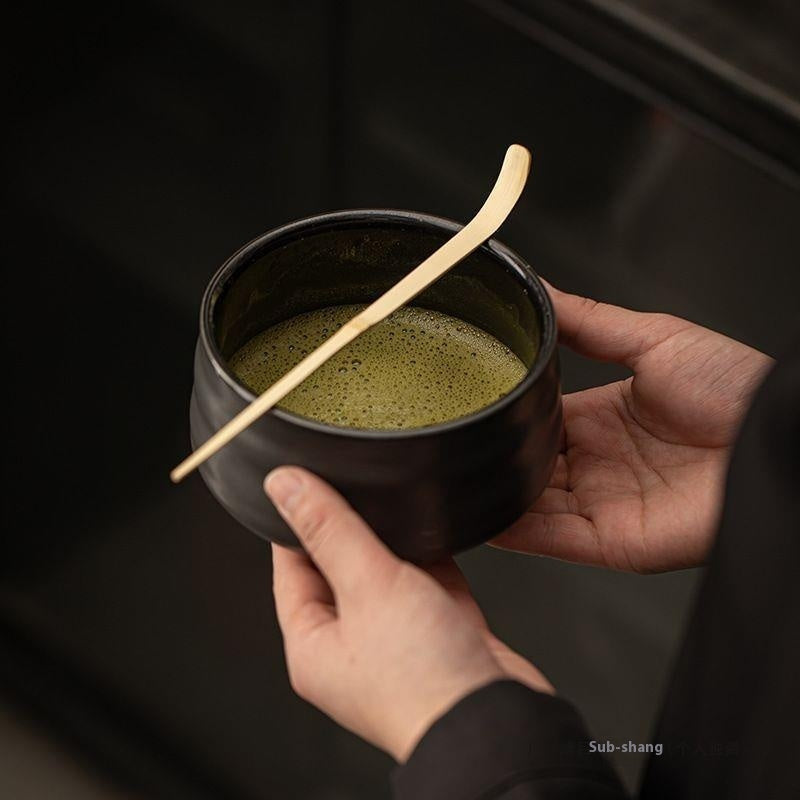 Hands whisking green matcha in wave pattern black chawan bowl